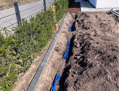 Geöffneter Graben entlang eines Hauses mit verlegten blauen Rohren und neben einem Zaun stehen grüne Büsche.