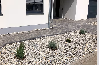 Vorgarten mit kleinen Kieselsteinen und vereinzelten Sträuchern vor einem modernen weißen Haus.