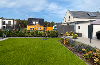 Garten mit gepflegtem grünem Rasen, Blumenbeeten und modernen Häusern im Hintergrund unter blauem Himmel.