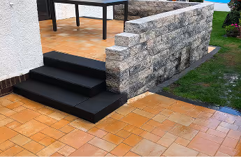 Schwarze Treppe neben einer hellgrauen Steinmauer führt zu einer Terrasse mit terrakottafarbenen Fliesen, angrenzend an einen Rasen.