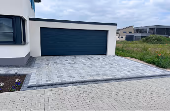 Einfahrt und geschlossene Garage eines modernen Hauses mit gepflastertem Boden und einer Wiese rechts.