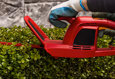 Person schneidet mit einer roten Akku-Heckenschere eine grüne Hecke.