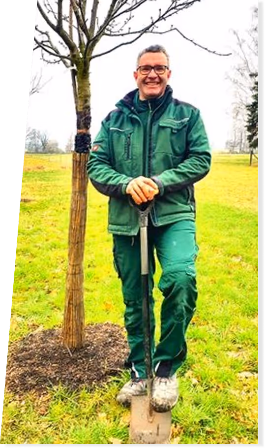 Mann in grüner Arbeitskleidung steht lächelnd auf einer Schaufel neben einem frisch gepflanzten Baum auf einer grünen Wiese.
