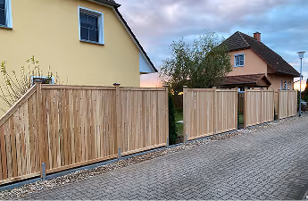 Holzzaun mit vertikalen Brettern entlang eines Gehwegs vor zwei Häusern bei Sonnenuntergang.
