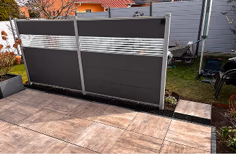 Moderner Sichtschutz aus dunklen Paneelen mit horizontalem Streifen auf einem gepflasterten Gartenweg.