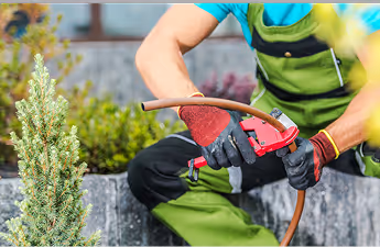 Person in grüner Arbeitskleidung und Handschuhen repariert einen Gartenschlauch mit einem Werkzeug vor einer Steinmauer.