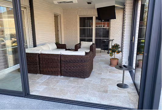 Überdachte Terrasse mit braunem Rattan-Sofa, kleinem runden Tisch, Topfpflanze mit Orangen und einem an der Wand montierten Fernseher.
